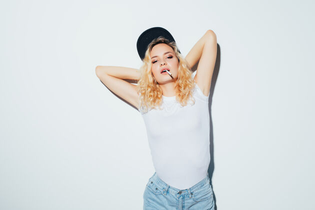 女孩一个年轻的金发女人的肖像 她抽烟 穿着白色t恤 戴着帽子 孤立地站在白色的墙上休闲时尚华丽
