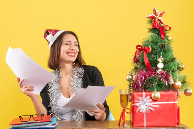 时尚穿着西装 戴着圣诞老人帽 戴着新年装饰品 手里拿着文件 坐在一张桌子旁 桌子上放着圣诞树圣诞老人坐着文件