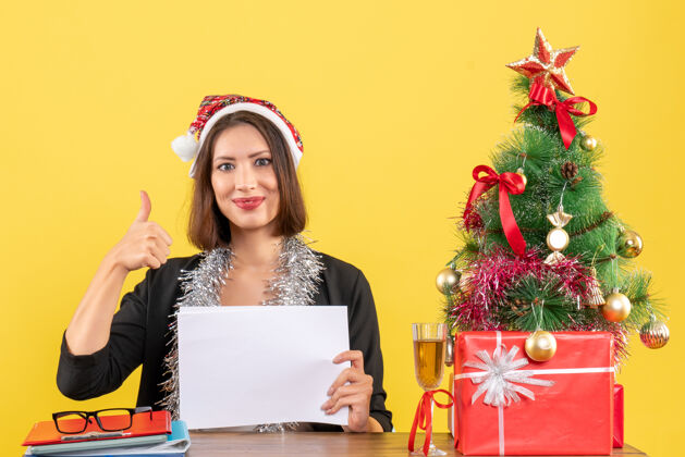 西装一位身着西装 戴着圣诞老人帽 戴着新年装饰品 面带微笑的商务女士独自一人做着“ok”手势 坐在办公室里一张桌子旁 桌上放着圣诞树好快乐工作