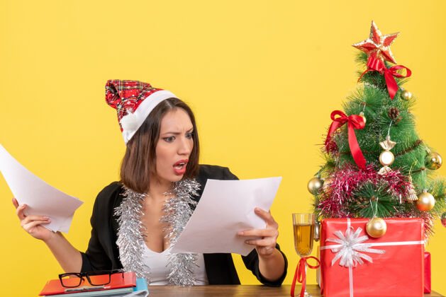 树穿着西装 戴着圣诞老人帽 戴着新年装饰品 情绪激动的商务女士正在检查文件 坐在一张桌子旁 桌子上放着圣诞树文件封面女郎年