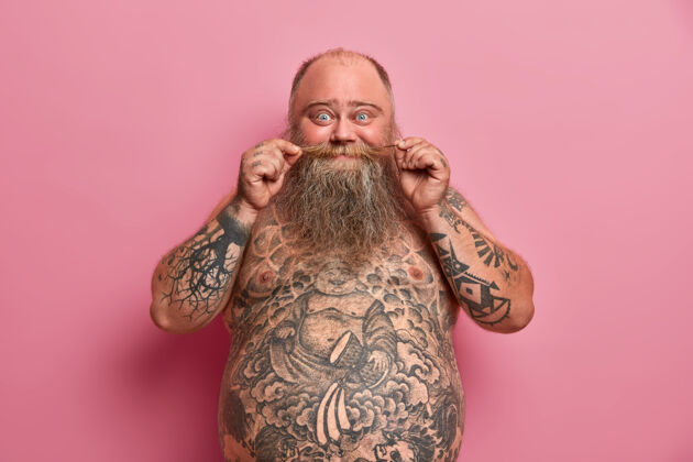 室内搞笑的胡须男人摸胡子 大肚子赤裸着站着 纹身 和朋友聊天 对着粉色的墙摆姿势赤膊上阵的肥胖男人人 营养 体型大站立脂肪