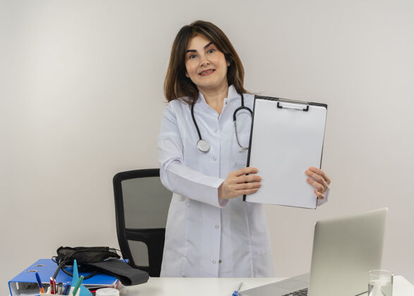 抱着微笑的中年女医生穿着医用长袍和听诊器坐在办公桌旁 手里拿着医疗工具和笔记本电脑成人坐着办公桌