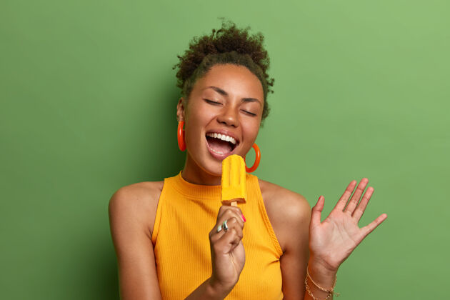 姿势快乐无忧无虑的非洲裔美国妇女像在麦克风里唱着黄色的冰淇淋 享受着美味的夏日产品 非常快乐 隔着鲜艳的绿色墙壁 举手 露出洁白的牙齿新鲜休闲大笑