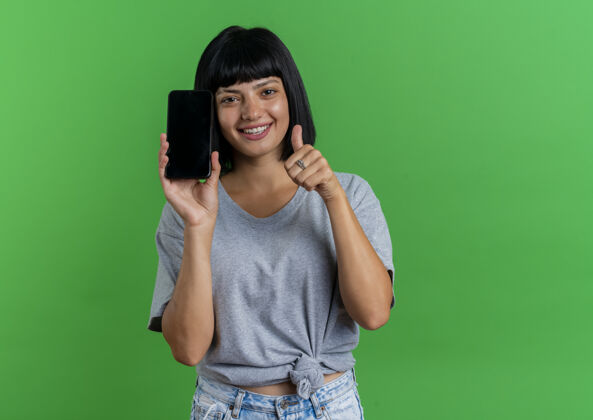 微笑微笑着的年轻黑发白人女孩手持手机 竖起大拇指 在绿色背景上与复印空间隔离开来拇指年轻向上