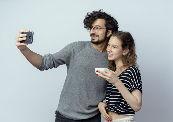 站立一对年轻的男女 快乐的男人用他的智能手机拍下了他们的照片 而他的女朋友站在他旁边喝着白色背景的咖啡使用情侣手机