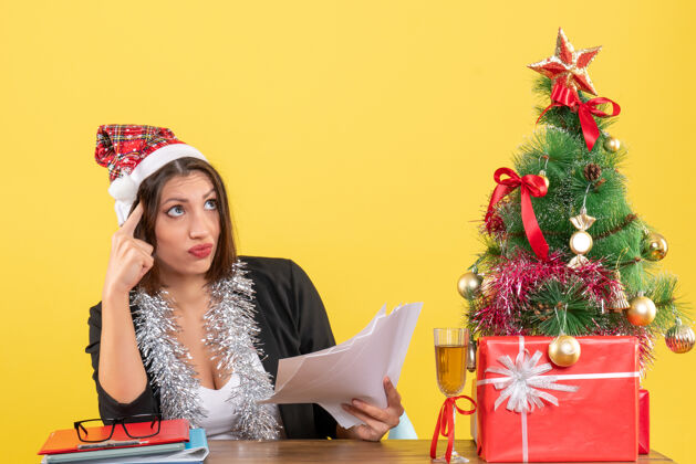 圣诞老人困惑的商务女士穿着西装 戴着圣诞老人的帽子和新年装饰品 手里拿着文件 坐在一张桌子旁 桌子上放着圣诞树桌子文件坐着