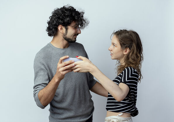 照片一对年轻漂亮的夫妇手持智能手机看着对方 在白色的背景上合影留念抱着每个人拿着