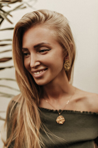 女人快乐快乐的长发金发棕褐色女人 穿着深绿色t恤 戴着金色珠宝 真诚地微笑着 在外面的白墙附近摆姿势成人优雅金发