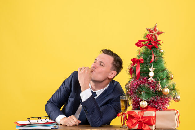 坐着正面图幸福的男人坐在圣诞树旁的桌子上做着雪夫亲吻的手势 黄色背景上的礼物手势幸福的人衣服