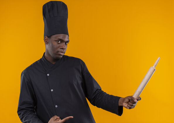 点年轻自信的非洲裔美国厨师穿着厨师制服 拿着并指着橘黄色背景上的独立擀面杖厨师别针制服