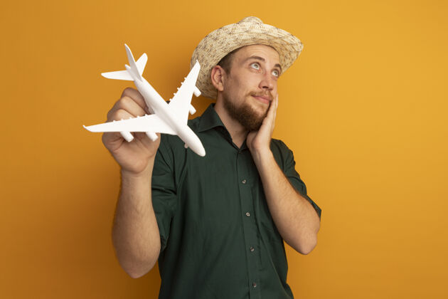 模特戴着沙滩帽的金发帅哥高兴地把手放在脸上 把飞机模型孤立地放在橙色的墙上帽子海滩脸