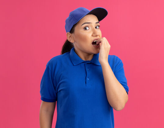 帽子身穿蓝色制服 头戴鸭舌帽的年轻女送货员站在粉红色的墙上 看着前面紧张紧张 咬牙切齿的指甲压力站立咬人