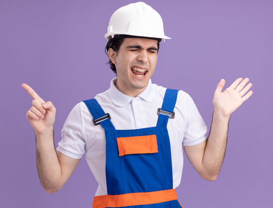 头盔年轻的建筑工人穿着建筑制服 戴着安全帽 兴高采烈地用食指指着站在紫色墙上的一边年轻人站安全