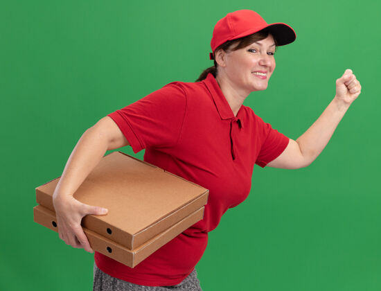 送货身着红色制服 头戴鸭舌帽的快乐女送货员正在绿色墙壁上为顾客运送比萨饼盒送货跑步快乐