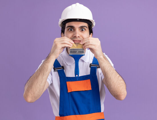 刷子身穿施工制服 头戴安全帽 手持油漆刷 模仿胡须的年轻建筑工人站在紫色的墙壁上 在工作中享受乐趣男人胡须工作
