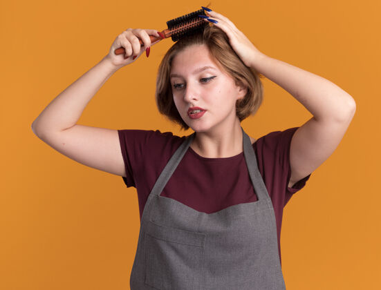 脸年轻漂亮的女理发师 围着围裙 用梳子梳头 严肃的脸站在橘色的墙上年轻女人理发师