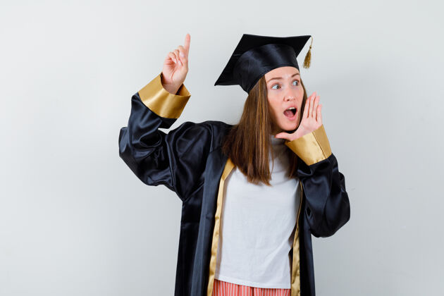 微笑毕业的女人指着头 穿着休闲服 手靠近张开的嘴 穿着制服 看上去很惊讶保持大学年轻