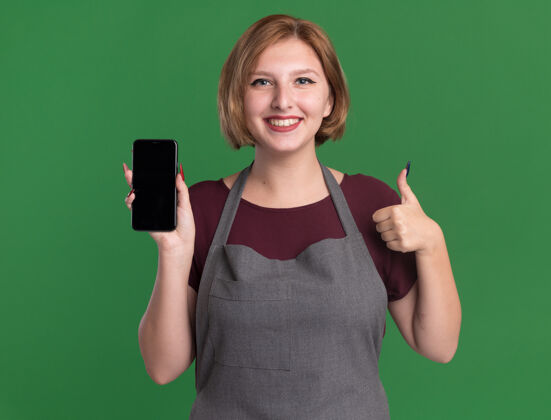 拇指年轻漂亮的女理发师 围着围裙 拿着智能手机 站在绿色的墙上 面带微笑 竖起大拇指女人漂亮围裙
