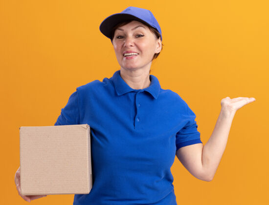 胳膊身穿蓝色制服 戴着帽子的中年送货妇女举着纸板箱 手举东西 面带微笑地看着前面站在橙色的墙上微笑介绍中间