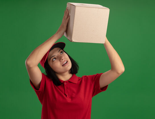 立场身穿红色制服 头戴鸭舌帽的年轻送货女 头上顶着一个纸板箱 站在绿色的墙上饶有兴趣地看着它头兴趣年轻