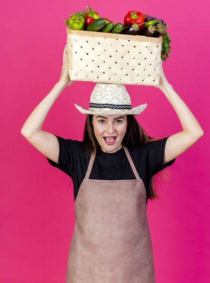 头快乐美丽的园丁女孩穿着制服戴着园艺帽把蔬菜篮子放在头上隔离在粉红色的背景上园艺制服花园