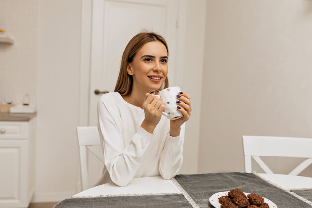 欢笑在厨房喝咖啡的无忧无虑的女孩穿着白衬衫的愉快微笑的女人享受咖啡休息的照片人网络咖啡杯