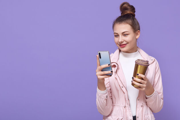女士开朗的女孩喝咖啡休息 手里拿着小玩意站着 阅读手机上的通知 更新最喜欢的应用程序 在看屏幕的时候输入信息和微笑 穿夹克 复制空间女孩1女士