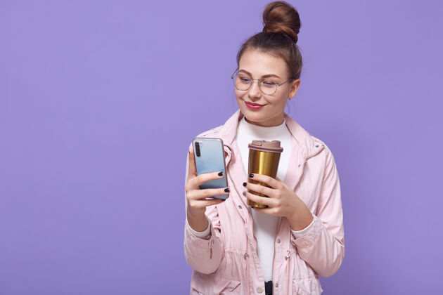 消息漂亮的女性戴着眼镜 穿着淡玫瑰色的夹克 隔着淡紫色的墙 喝着热马克杯里的热饮料 手里拿着智能手机 浏览着互联网移动模型消息