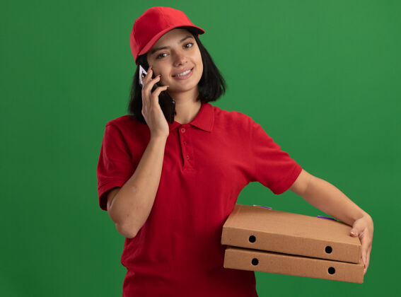 女孩身穿红色制服 头戴鸭舌帽 拿着比萨饼盒的年轻送货员站在绿色的墙上微笑着用手机交谈站着拿着电话