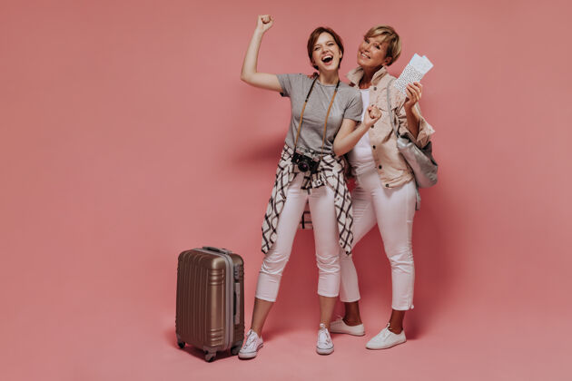 白色快乐的两个短发女人 穿着白色紧身裤和运动鞋 微笑着 拿着手提箱 相机 票和包在孤立的背景上摆姿势成人游客衬衫