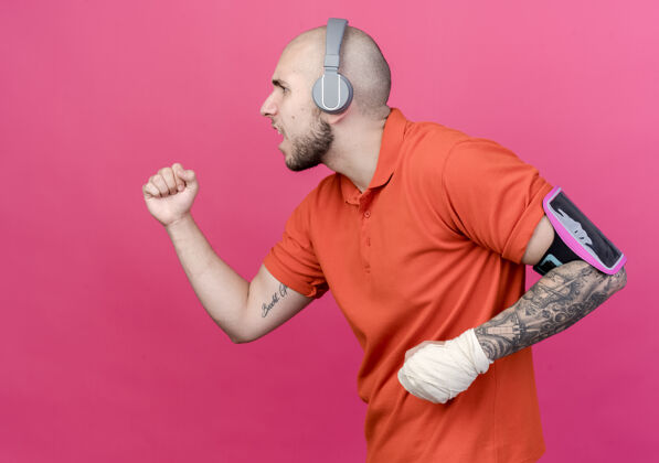 假装在侧视图中 年轻的运动型男子 手腕缠着绷带 头戴耳机 胳膊上戴着电话带 假装在粉色背景下孤立地奔跑跑步运动手腕