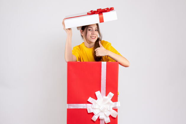 帽子前视图兴高采烈的女孩与圣诞老人帽子制作拇指向上的标志站在大圣诞礼物后面大使圣诞节
