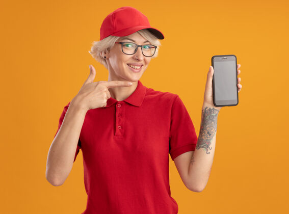 女士身着红色制服 头戴眼镜 戴着智能手机 食指指着智能手机 站在橙色墙壁上开心地微笑的年轻送货员欢呼穿着指点