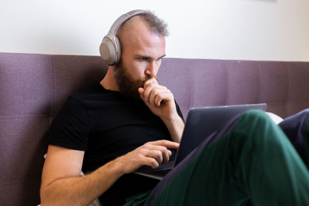房间一个留着大胡子的白种人戴着耳机在卧室的床上工作 在家里用笔记本电脑 打字 思考早晨在线舒适