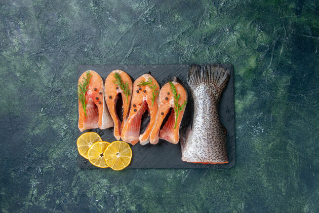 混合新鲜生鱼片 青椒和柠檬片的俯视图 放在蓝黑混合色桌上的深色托盘上食物切片托盘