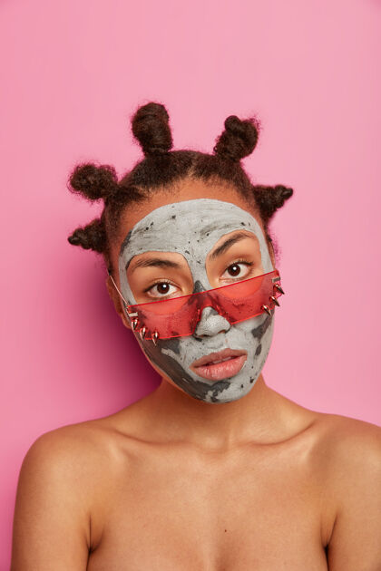 清新严肃冷静的女人戴着美容面具 赤裸上身站在室内 戴着玫瑰色太阳镜 减少皱纹和黑头 隔离在粉红色的墙上垂直拍摄年轻 健康美容身体姿势
