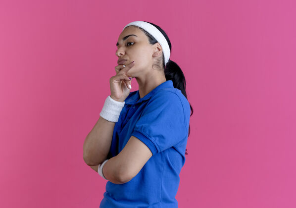 年轻年轻体贴的白人运动女性戴着头带和腕带站在一边 把手放在下巴上的粉红色与复制空间侧头带下巴