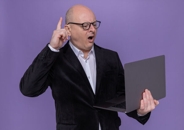 手指中年光头西装革履 戴着眼镜 拿着笔记本电脑 惊讶地伸出食指 站在紫色的墙上 有了新的想法穿目录西装