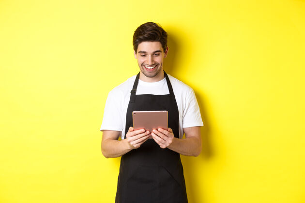 模特穿着黑色围裙的卖家看着数码平板电脑屏幕 微笑着高兴 站在黄色背景上工作室成人帅哥