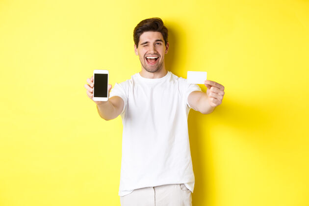 站着快乐的男人在手机屏幕上展示好的在线报价 拿着信用卡眨眼 站在黄色背景上网购手机成人