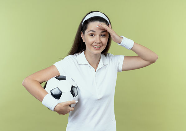 女人戴着头巾拿着足球的年轻健身女士站在轻质墙壁上 看起来很困惑 因为她站错了足球光头