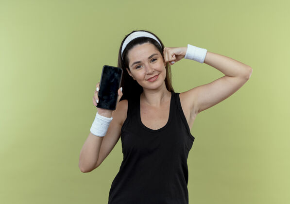 二头肌戴着头巾的年轻健身女士展示了智能手机举起的拳头 展示了站在轻质墙上自信的二头肌站立移动信心