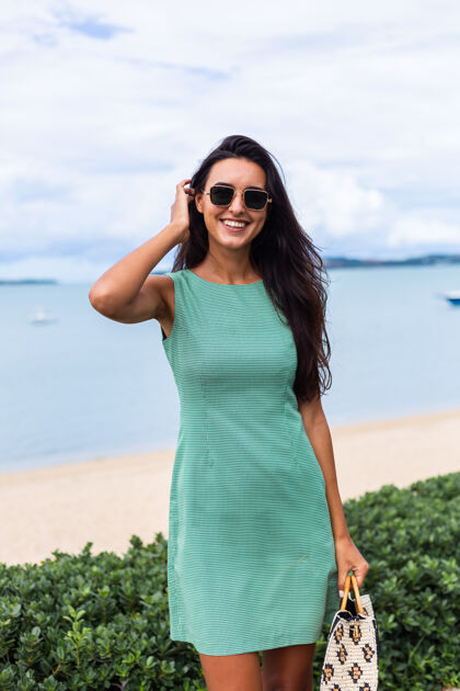女人漂亮时尚的快乐女人 穿着绿色夏装 带着包 度假时戴着太阳镜 背景是蓝色的大海旅游华丽度假