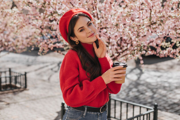 花园穿着红色毛衣 戴着红帽子的年轻女子手里拿着一杯硬纸板咖啡年轻女子拿着茶杯 对着盛开的樱花真诚地微笑着太阳镜芳香欢呼