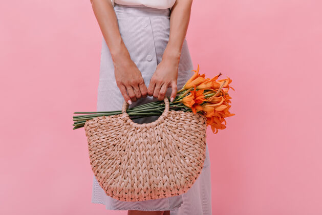 梦幻女士带着编织袋和粉色背景上的花朵摆出姿势身着蓝色浅色裙子的女士拿着一个带有橙色美丽花束的草包卷发深色室内