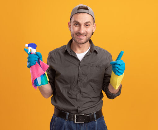 男人年轻的清洁工穿着休闲服 戴着橡胶手套 戴着帽子 手里拿着喷雾瓶和抹布 面带微笑 站在橘色的墙上竖起大拇指休闲破布拇指
