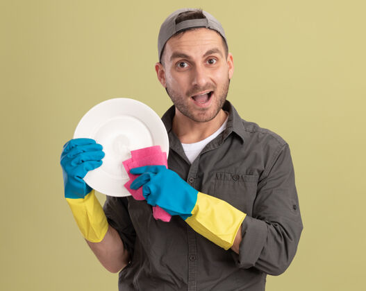 清洁年轻的清洁工穿着休闲服 戴着橡胶手套 手里拿着盘子和抹布 站在绿色的墙边 看上去很开心 很兴奋盘子橡胶破布