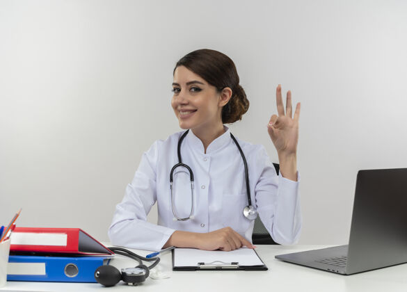 可以面带微笑的年轻女医生穿着医用长袍 听诊器坐在办公桌旁 带着医疗工具和笔记本电脑 在白色的墙上贴着“一切正常”的牌子医生听诊器坐着