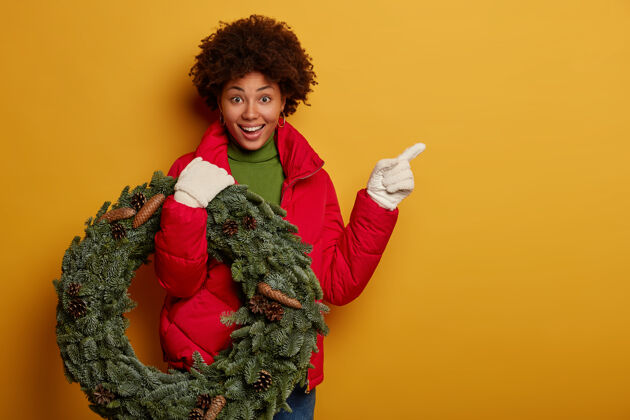 微笑快乐的非洲女人向她家走去 穿着红外套 戴着白手套 拿着圣诞花环 指着空白处 站在黄色的背景下人满意季节
