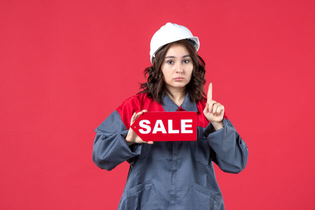 销售正面近距离观看穿着制服 戴着安全帽的紧张的女工人展示销售图标 并指着隔离的红墙人肖像前面